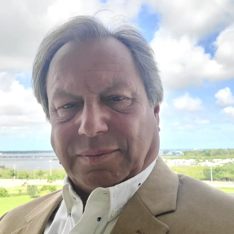 Man in beige blazer takes a selfie with a scenic outdoor backdrop.